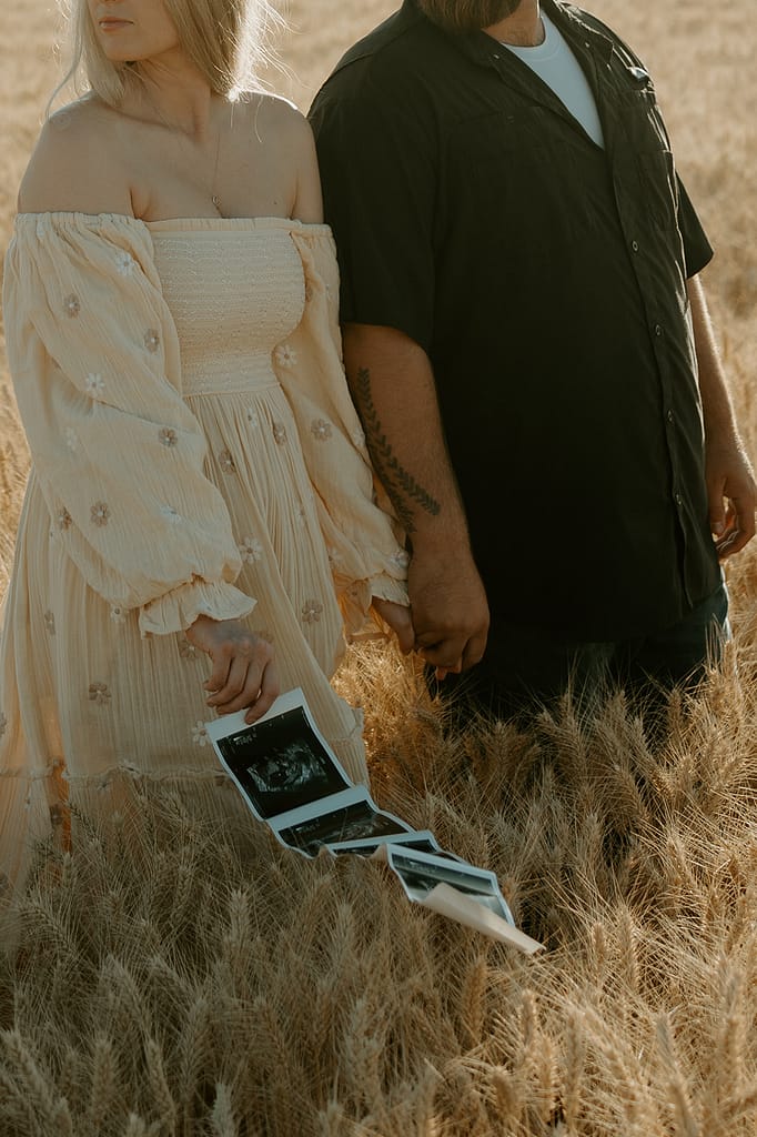 Pregnancy reveal photo shoot in a wheat field: Haley and Tate stand together amidst golden wheat stalks, holding ultrasound photos and a tiny onesie.