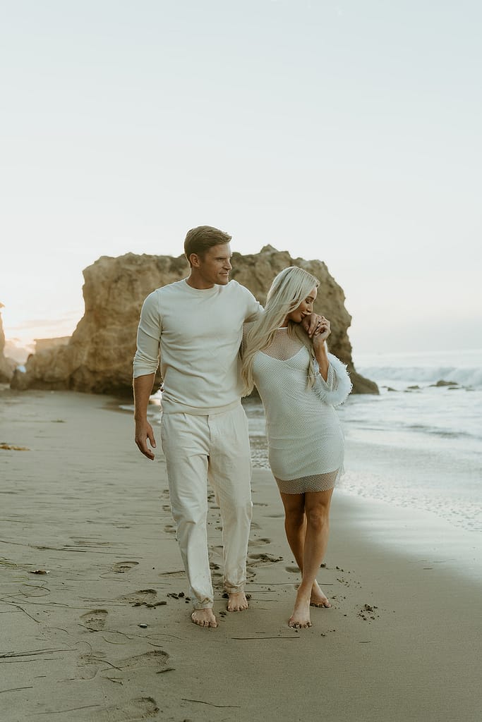 Engaged couple walking along the sandy shore of Malibu Beach during sunrise.