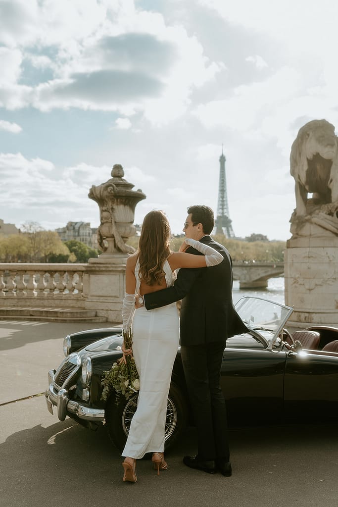 A couple poses near a vintage car parked in front of the Eiffel Tower in Paris, France, capturing a timeless and romantic moment in the city of love.