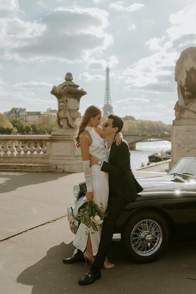 A couple poses on a vintage car parked in front of the Eiffel Tower in Paris, France, capturing a timeless and romantic moment in the city of love.