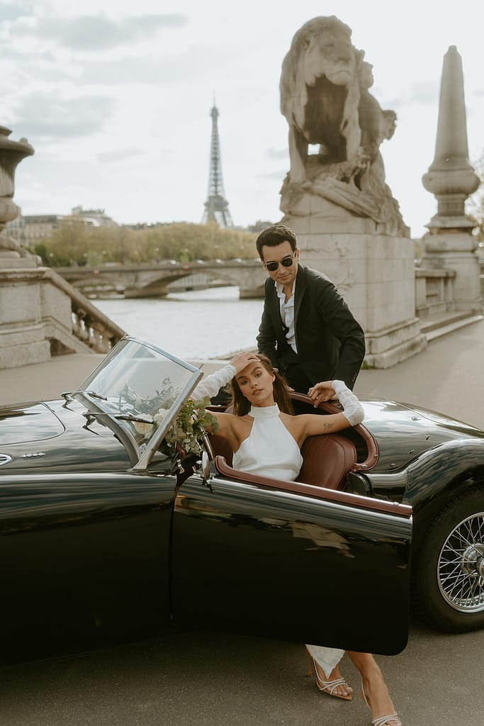 A couple poses in a vintage car parked in front of the Eiffel Tower in Paris, France, capturing a timeless and romantic moment in the city of love.