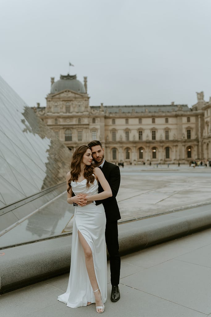 A couple embraces in front of the iconic Louvre Museum in Paris, France, with ornate architecture in the background.